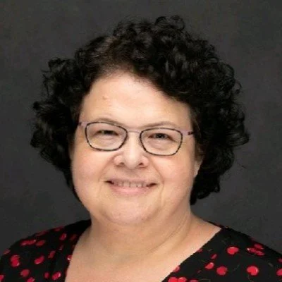 A woman with curly dark hair wearing glasses and a black top with red polka dots, smiling at the camera against a dark background.